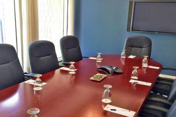 The image shows a conference room with a long table, office chairs, a speakerphone, upside-down glasses, a tray of candy, and a mounted TV screen.