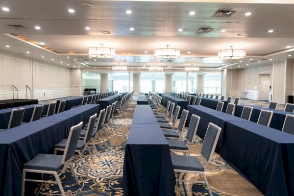 A large, well-lit conference room with long tables covered in blue tablecloths and rows of chairs arranged in parallel, facing a stage.