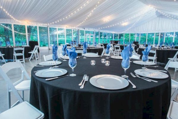 The image shows an elegantly decorated event tent with round tables covered in black tablecloths, blue napkins in wine glasses, and white chairs.