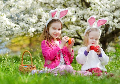 Two children with bunny ears sit on grass holding Easter eggs near a basket, with blooming trees in the background.
