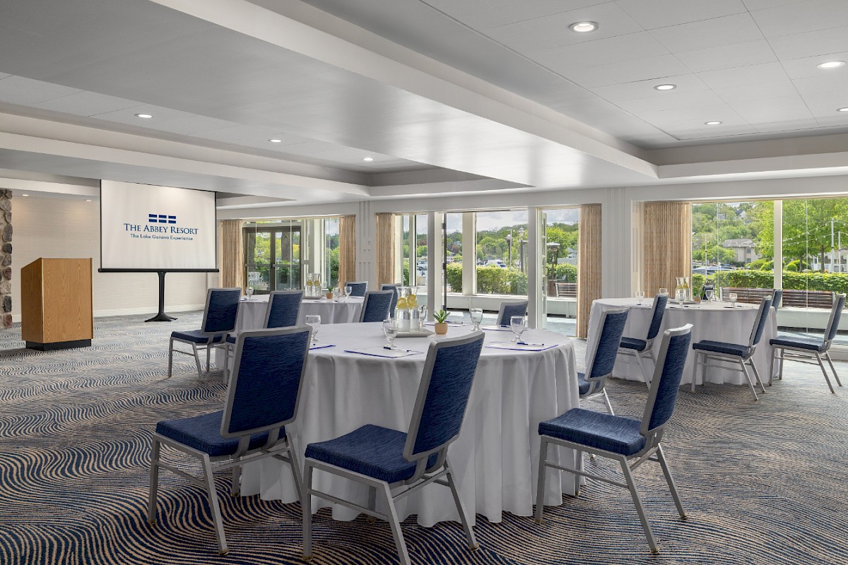 A conference or event room with round tables, blue chairs, a podium, and a large screen, arranged for a presentation or meeting at a hotel or venue.