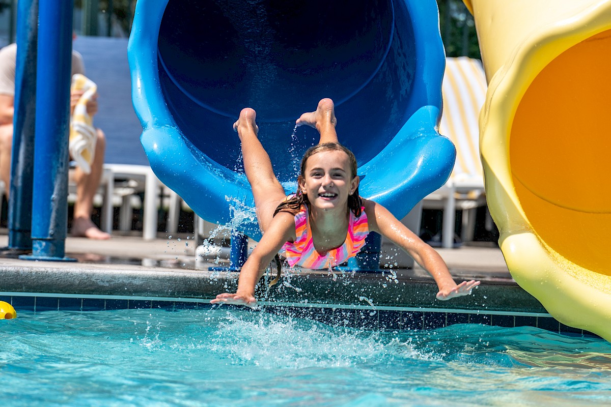 A girl emerges from a blue slide into the pool, smiling, during an outdoor summer day at a water park.