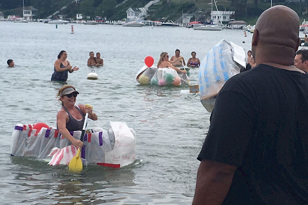 People are participating in a fun event in a lake. Some individuals are in makeshift rafts, while others are swimming or watching from the shore.