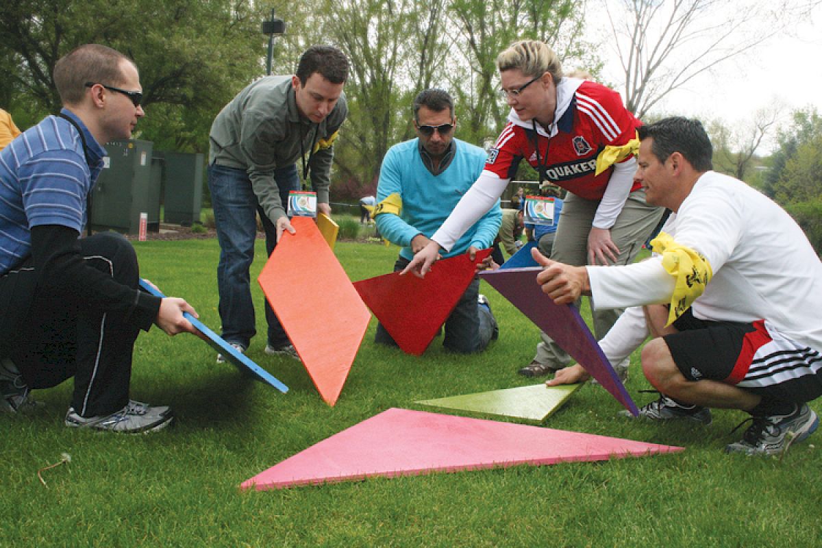 A group of five people outdoors are arranging colorful triangular tiles on the grass, suggesting a team-building activity.