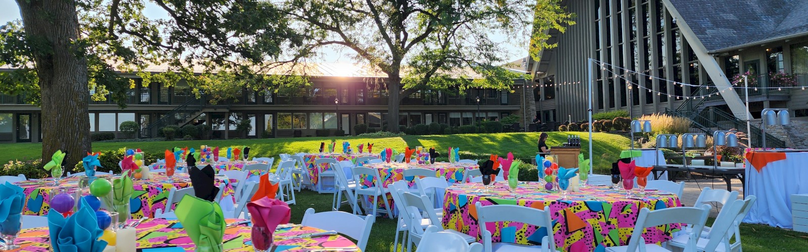 An outdoor event setup with colorful tablecloths and napkins, white chairs, and a building in the background, under sunny skies.