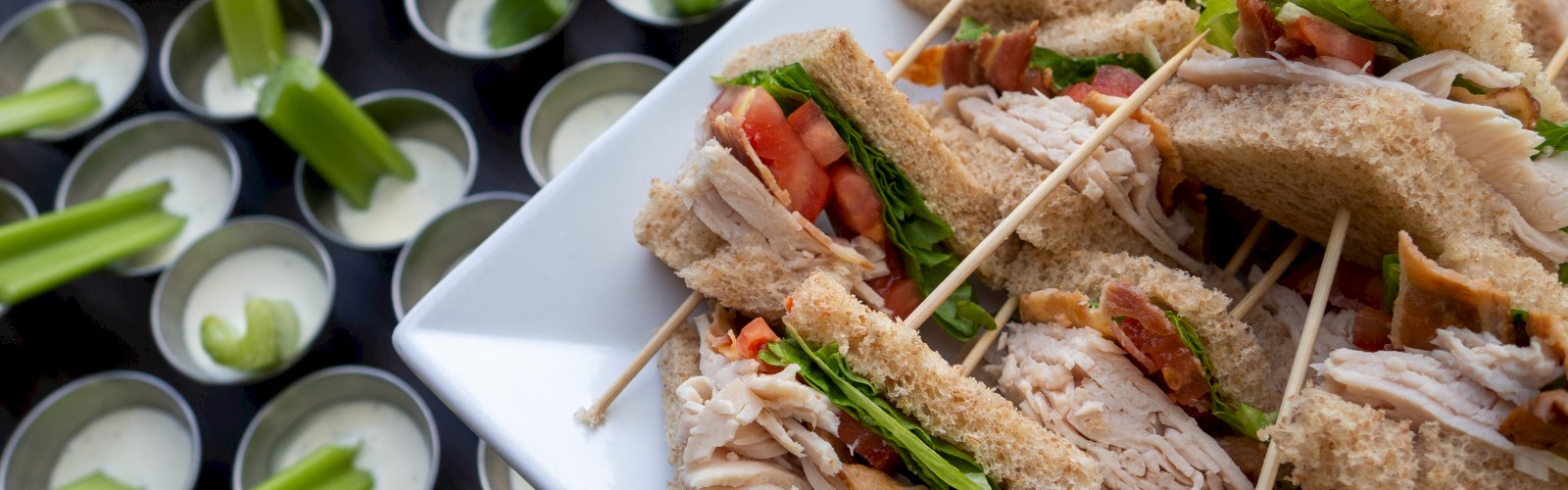 A platter of sandwiches with toothpicks, alongside cups of dipping sauce paired with celery sticks on a table, is shown in the image.