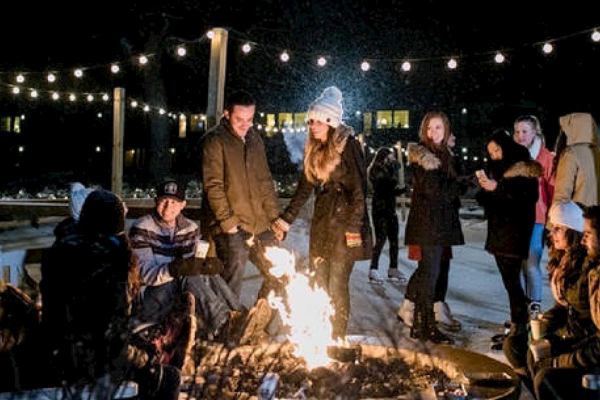 A group of people, dressed in winter clothing, are gathered around a fire pit outdoors at night, with string lights overhead.