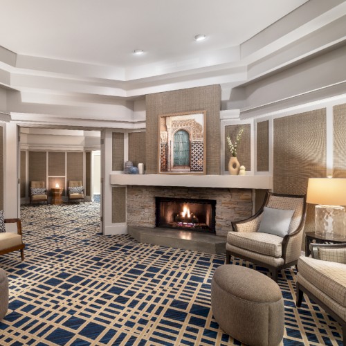 A stylish hotel lobby with a lit fireplace, plush seating, soft lamps, and a geometric carpet pattern, creating a cozy, upscale vibe.