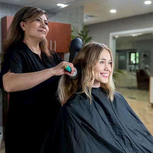 A hairdresser is brushing a client's blonde hair at a salon, both smiling and appearing happy in the brightly lit room.