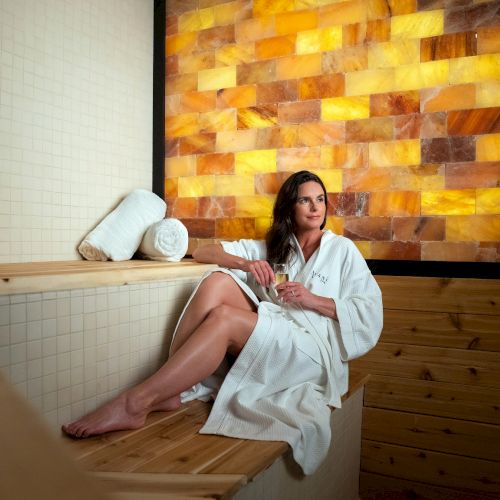 A person in a white robe is sitting on a wooden bench in a room with a warm backlit salt wall and rolled towels beside them.