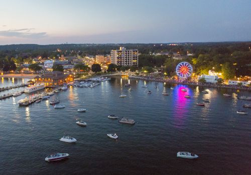 An aerial view of a waterfront with boats, a lit Ferris wheel, docks, and buildings in the evening. The skyline and horizon are visible.