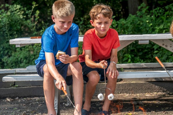 Two children are sitting by a fire pit, roasting marshmallows with sticks. They are outdoors with greenery in the background and a picnic table.