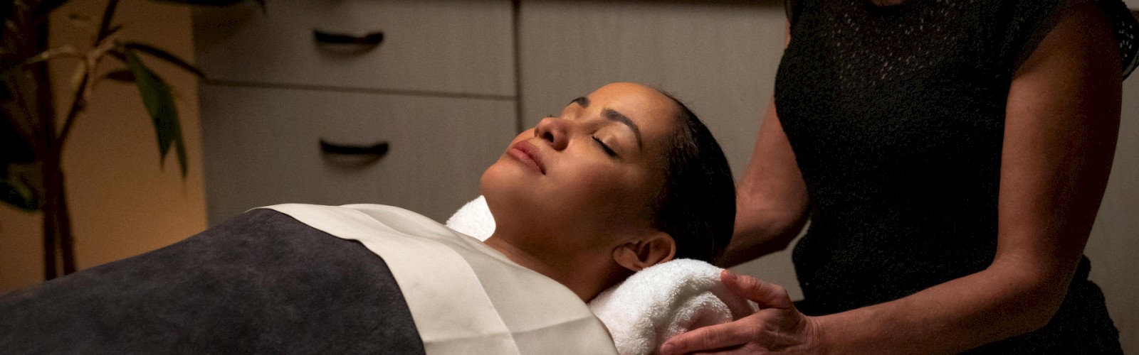 A woman is lying on a spa bed with a towel rolled under her neck, receiving a massage in a dimly lit room with candles.