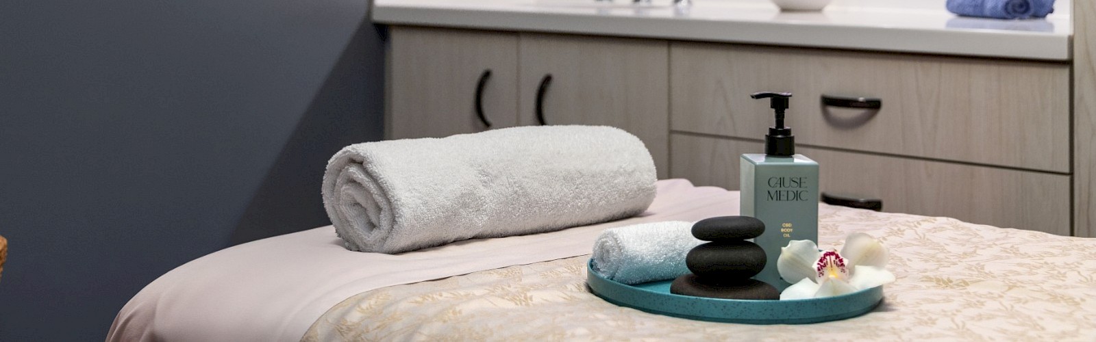 A cozy spa-like treatment room with a sleek vanity, soft lighting, a rolled towel and bamboo tray on a massage table, and calming decor.