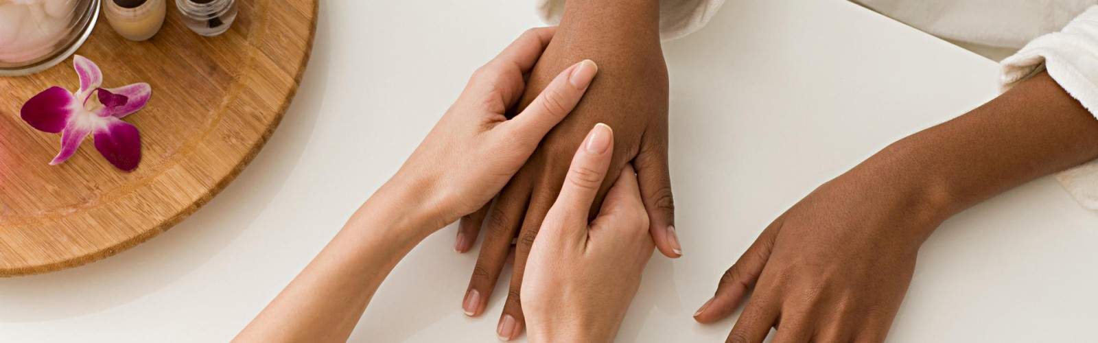 A person is receiving a manicure, with one individual's hands being held by another pair of hands, and manicure tools and an orchid on a wooden tray.