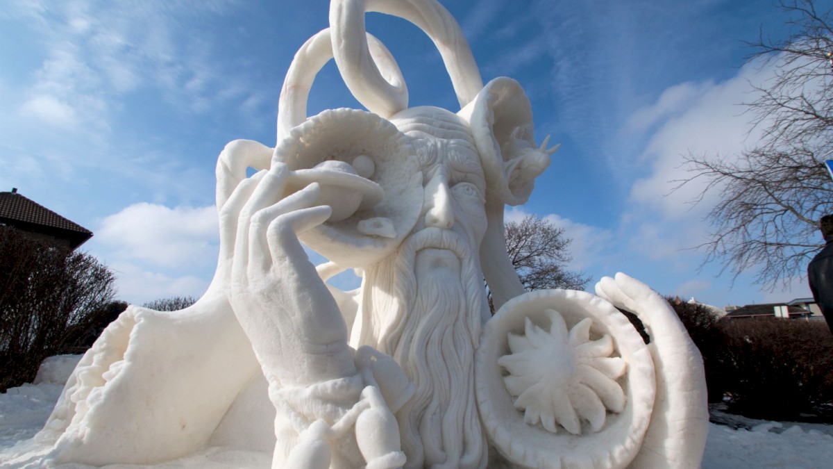 A large snow sculpture featuring a bearded man with elaborate, swirling hair and a seashell held to his ear, standing in a snowy outdoor setting.