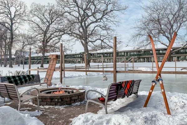 A snowy outdoor scene with a fire pit, benches, skating area, and crossed skis near leafless trees in winter.