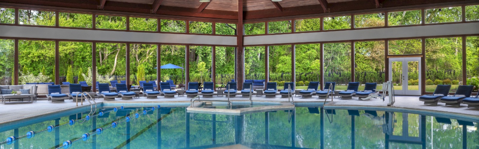 It's an indoor pool area with a hot tub, surrounded by lounge chairs under a wooden ceiling with large windows revealing a forest view.