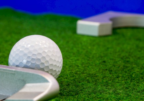 A golf ball is positioned on green turf near a club, with a flagstick and hole visible in the background.