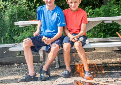 Two boys sit on a bench, roasting marshmallows over a campfire in a lush, green outdoor setting.