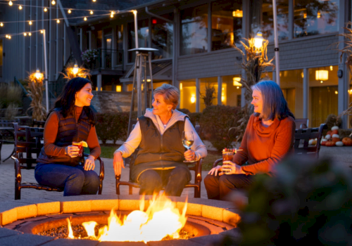Three people sit around a fire pit, enjoying drinks, with string lights and a cozy setting in the background.