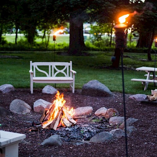 People are tending a campfire in a park-like setting with benches, torches, trees, and grassy areas.