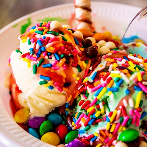A bowl of ice cream topped with colorful sprinkles, candies, and syrup with a spoon inside it.