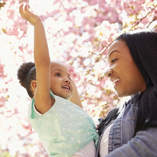 A woman holding a smiling girl under blooming pink cherry blossom trees.