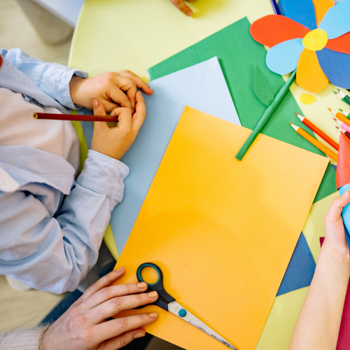 Children are making colorful paper crafts, including a rocket and a flower, using scissors, pencils, and paper on a yellow table.