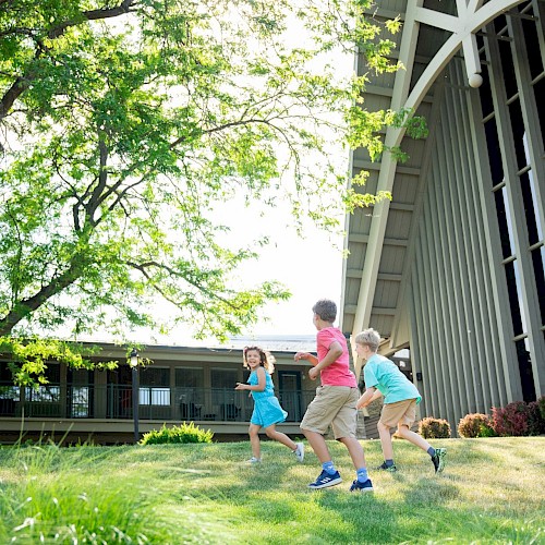 Three kids are playing and running on a grassy yard near a house, enjoying a sunny day in a lush, green outdoor setting.