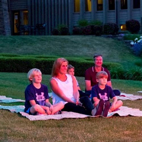 A family is sitting on blankets outdoors, watching an evening event on the grass in front of a residential area.