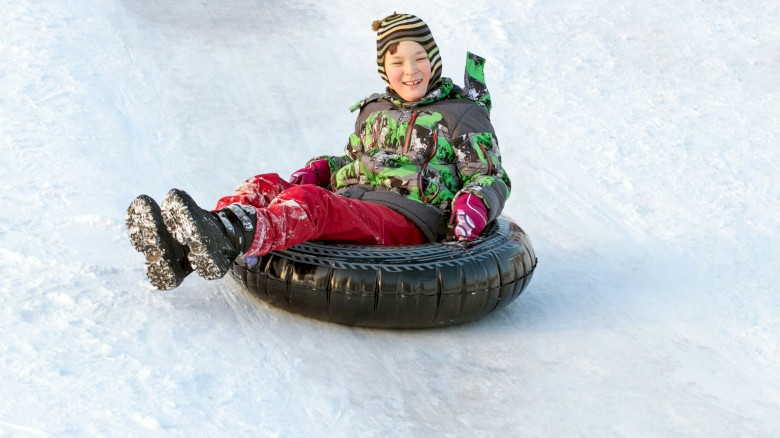 A child wearing colorful winter clothes is smiling while riding a snow tube down a snowy slope.