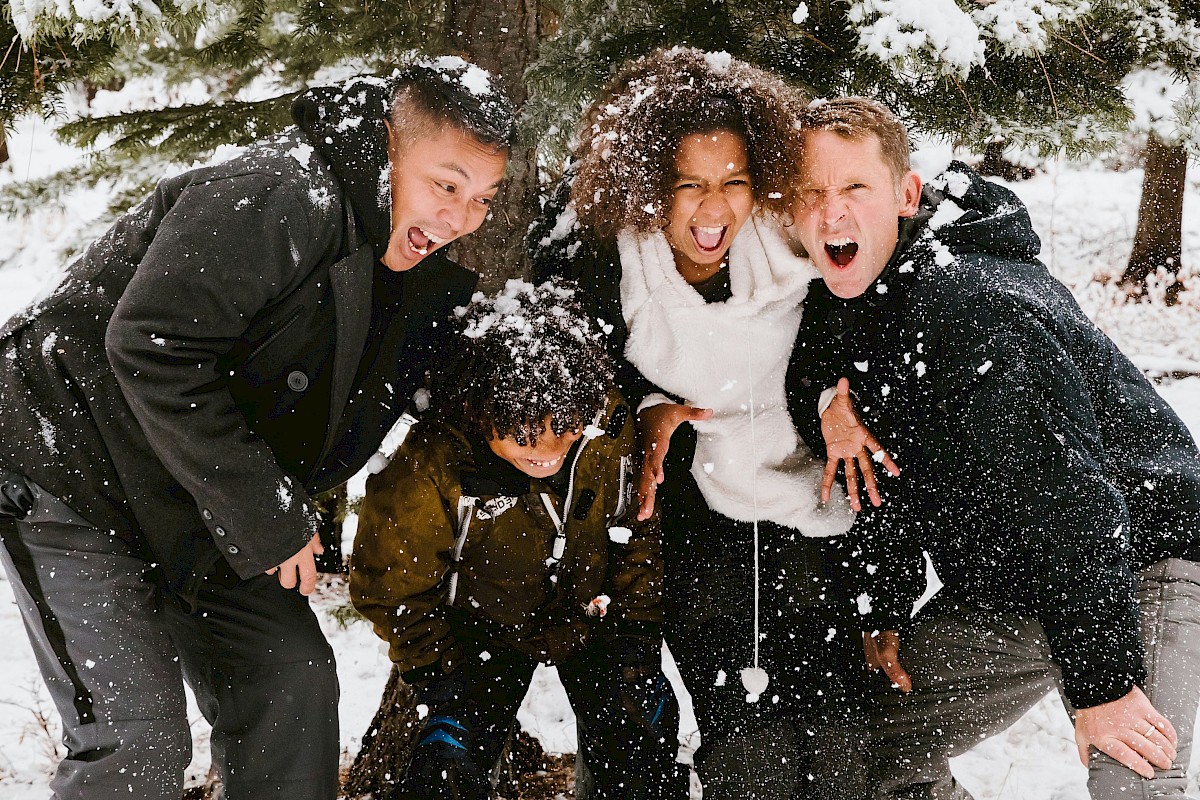 A group of five diverse friends happily playing in the snow under a tree, throwing snowballs and having fun outdoors.