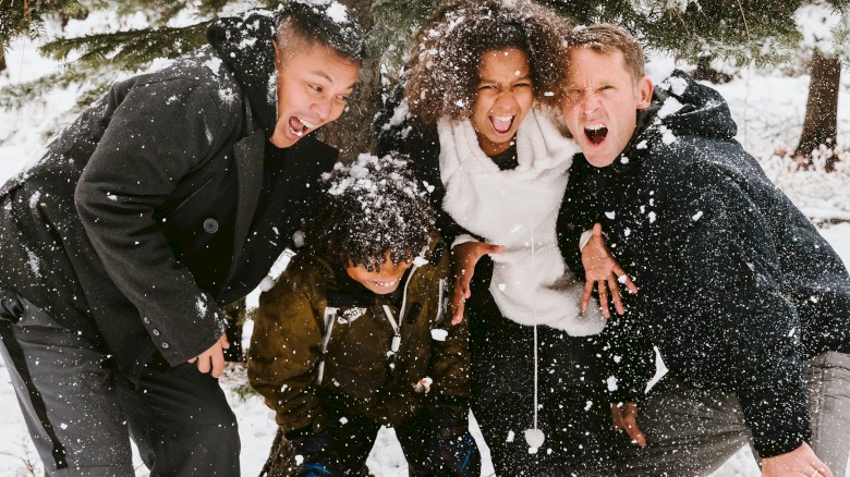 A group of five diverse friends happily playing in the snow under a tree, throwing snowballs and having fun outdoors.