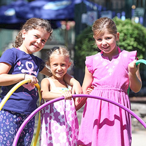 Three smiling young girls hold hula hoops and stand outdoors, enjoying a sunny day at a park or playground.