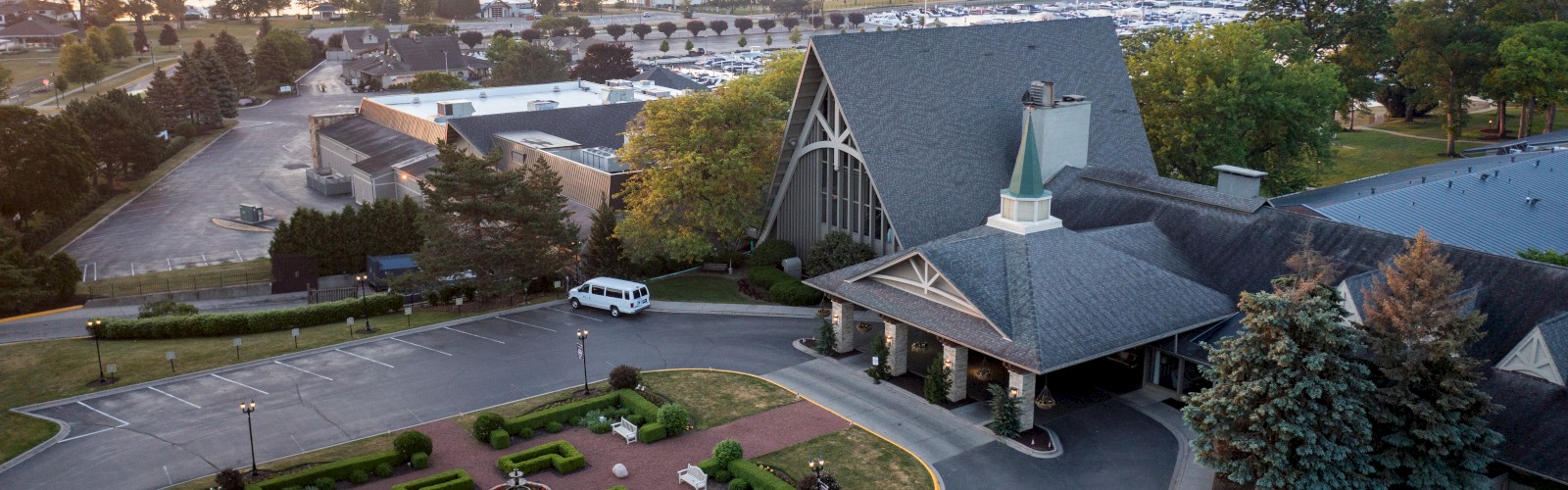 An aerial view shows a building with a manicured garden, nearby parking lot, and distant water body with surrounding greenery at sunset.