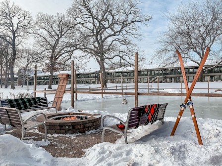 A snowy outdoor scene with a fire pit, benches, ice skating rink, and skis, surrounded by bare trees and a building in the background.