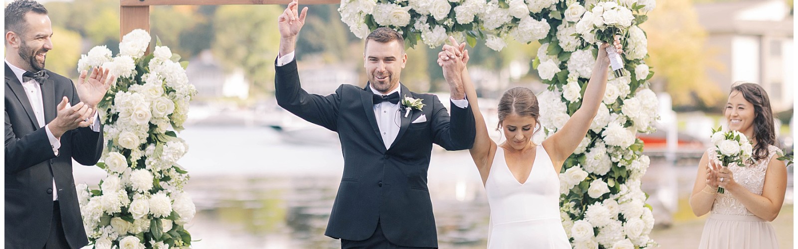 A newlywed couple celebrates under a floral arch with two people on each side applauding, situated outdoors by a lake or river.