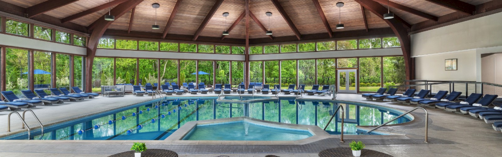 Indoor pool area with a large circular pool, surrounded by many lounge chairs and small tables with chairs; a high wooden, vaulted ceiling.