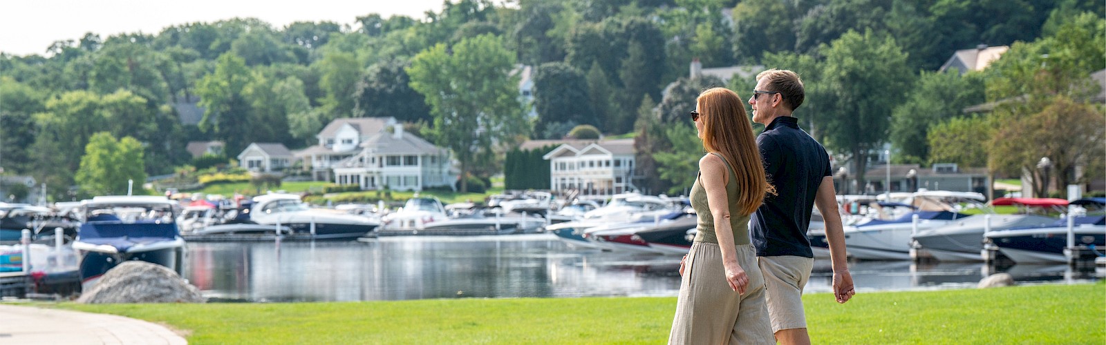 A couple walks along a park near a marina with boats and colorful houses in the background, enjoying a sunny day.