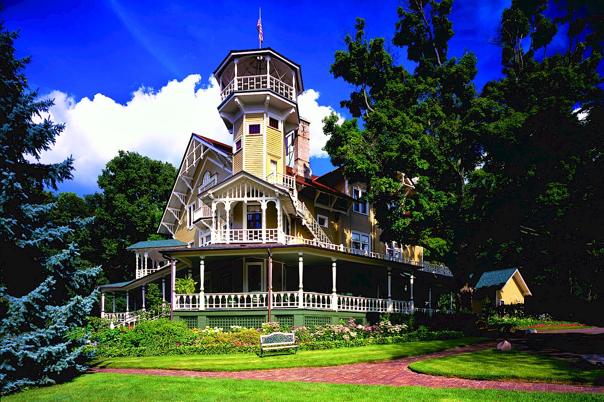 A large, ornate yellow Victorian house with a tower, surrounded by lush green trees and a well-kept garden on a bright day.