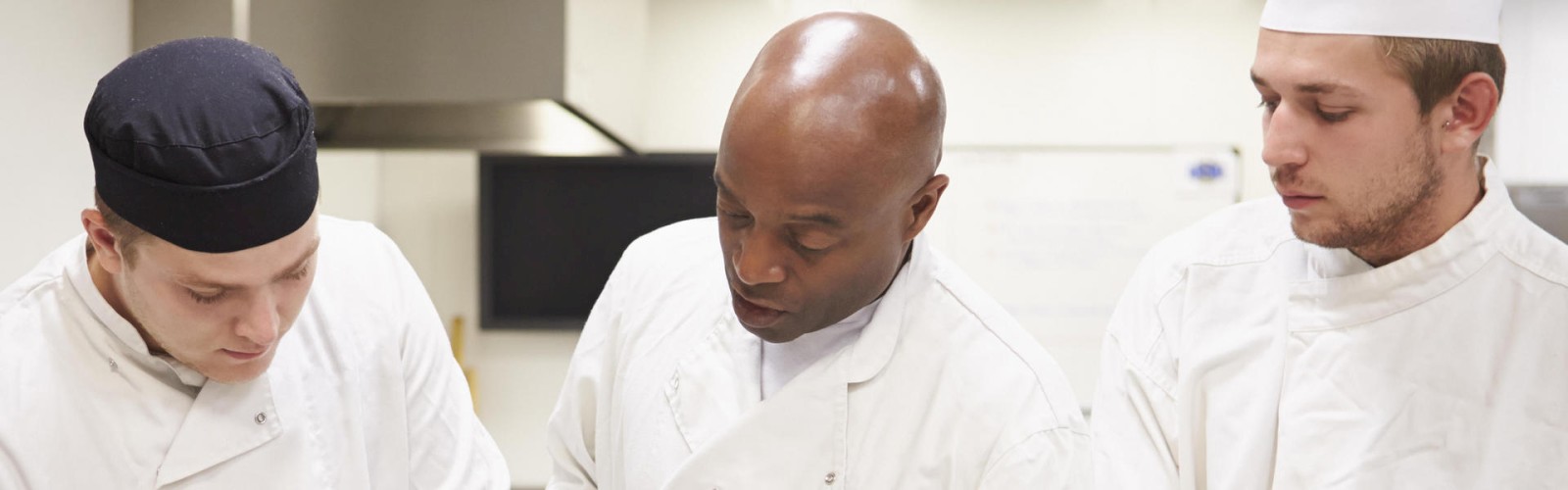 Three chefs in white uniforms are working and discussing something in a professional kitchen environment.