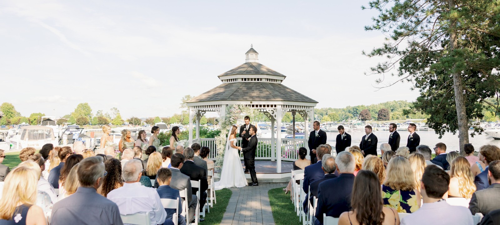 A couple is getting married under a gazebo by a waterfront, surrounded by guests and a wedding party, ending the sentence.