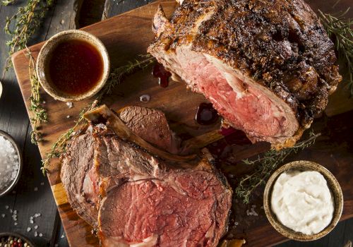 The image shows a cooked, sliced prime rib on a wooden board, accompanied by mashed potatoes, asparagus, a small cup of sauce, and garnishes.