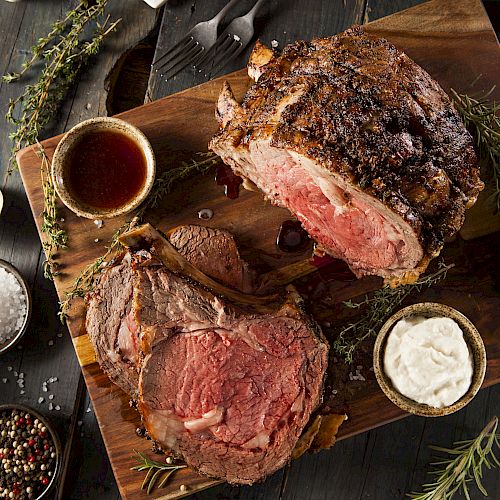 The image shows a cooked, sliced prime rib on a wooden board, accompanied by mashed potatoes, asparagus, a small cup of sauce, and garnishes.