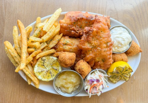 A plate of fish and chips with fries, lemon slices, sauces, coleslaw, and hush puppies on a wooden surface.