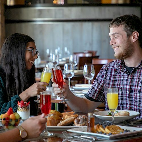 A group of people enjoying a meal at a restaurant, sharing a toast with drinks, surrounded by plates of food and desserts, and smiling warmly.