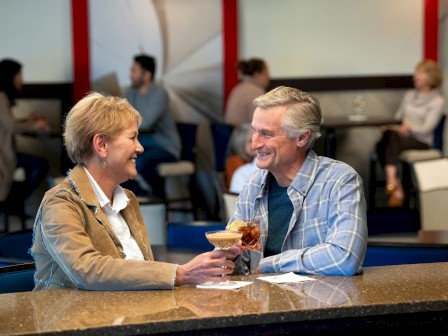 Two people are sitting at a bar, smiling, and holding drinks. Other patrons are in the background.