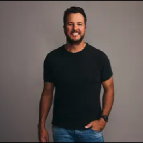 A smiling man in a black t-shirt and jeans stands against a neutral background, casually posing with one hand in his pocket.