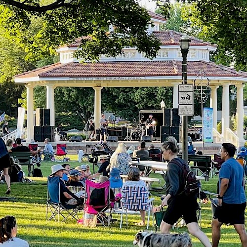A lively outdoor concert in a park with families, children, and musicians enjoying a sunny afternoon under green trees.